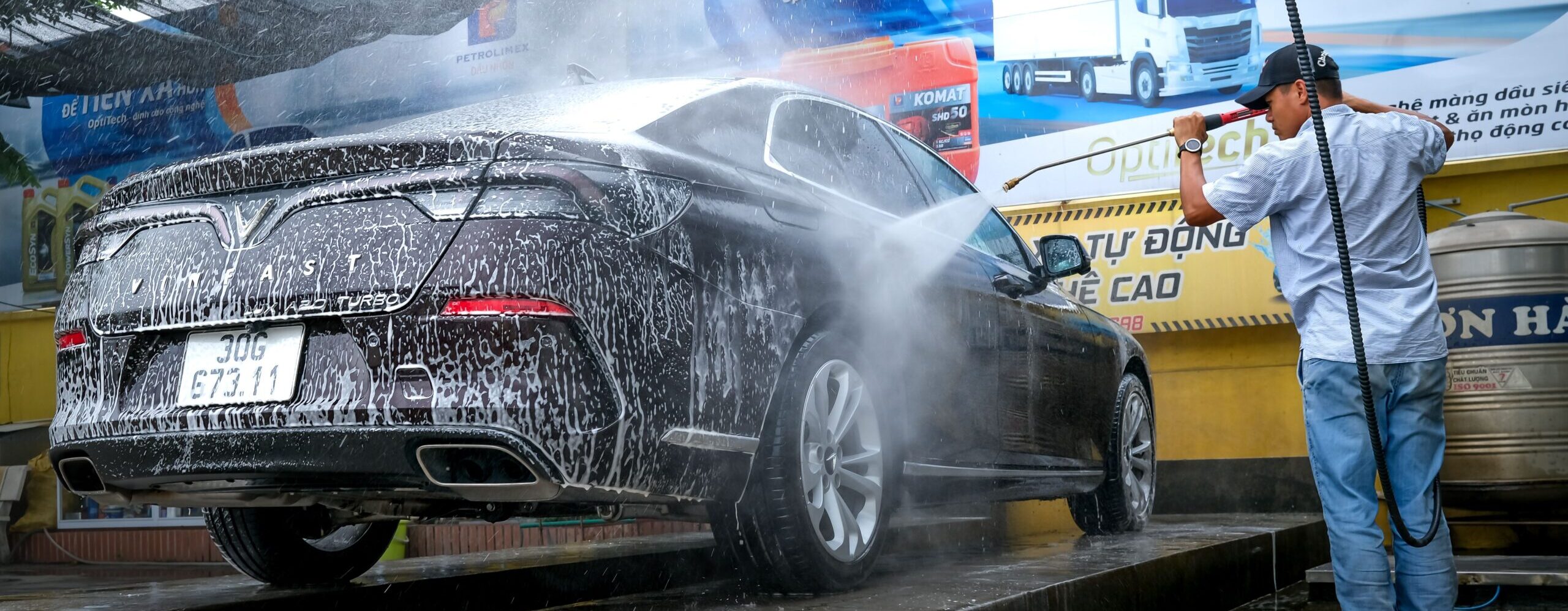 A black car is being cleaned with a power spray by a man at a car wash station outdoors.